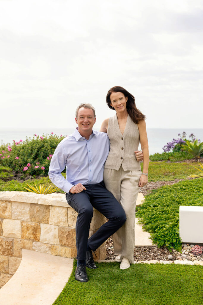 John Hood and Sally Hood, each with an arm around each other's side, standing in outdoor setting with flowers behind them.